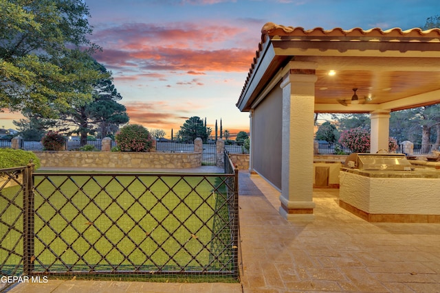 patio terrace at dusk featuring area for grilling, a ceiling fan, a gate, fence, and an outdoor kitchen