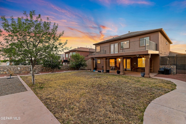 back of house at dusk with a patio area, fence, a balcony, a yard, and stucco siding