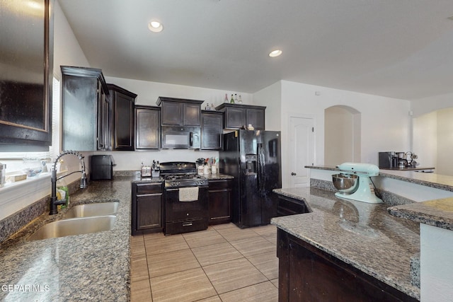 kitchen with dark stone countertops, a sink, arched walkways, dark brown cabinetry, and black appliances