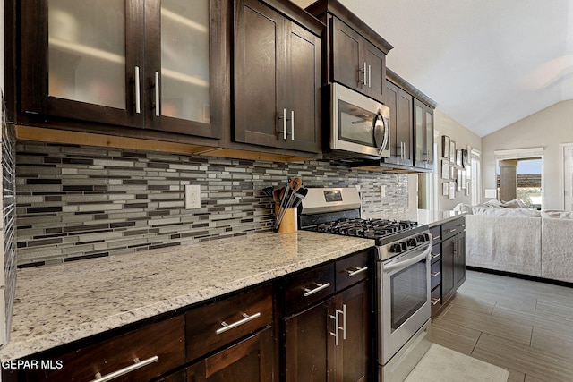 kitchen with lofted ceiling, dark brown cabinets, light stone counters, appliances with stainless steel finishes, and backsplash