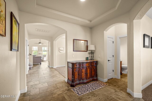 hallway with stone tile floors, arched walkways, a raised ceiling, baseboards, and visible vents