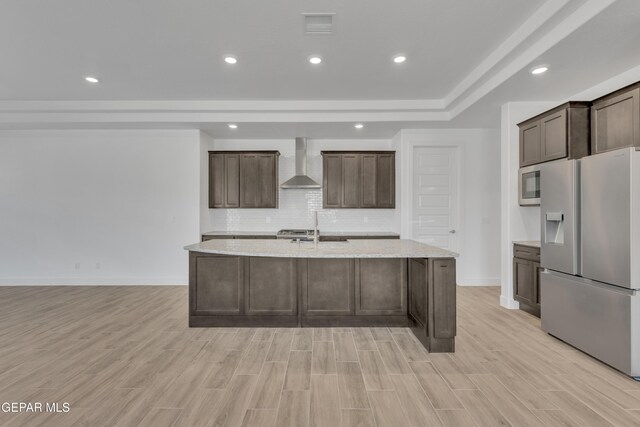 kitchen featuring wall chimney range hood, a sink, a kitchen island with sink, tasteful backsplash, and appliances with stainless steel finishes