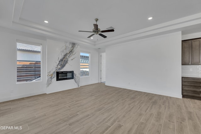 unfurnished living room with a ceiling fan, light wood-style floors, a premium fireplace, baseboards, and a raised ceiling