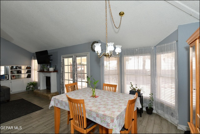 dining room with a notable chandelier, light wood-style flooring, a fireplace with raised hearth, a textured ceiling, and vaulted ceiling