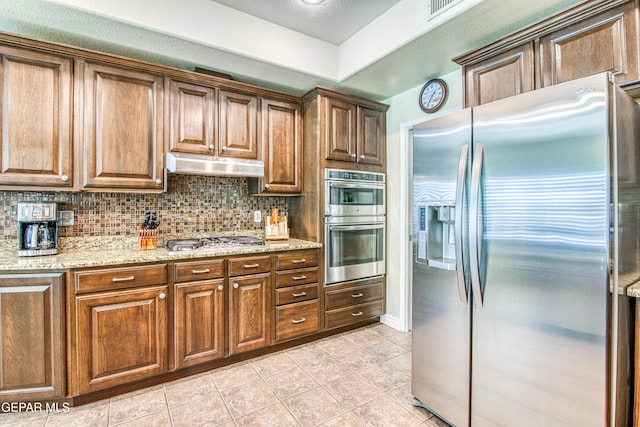 kitchen with tasteful backsplash, light tile patterned floors, light stone counters, under cabinet range hood, and stainless steel appliances