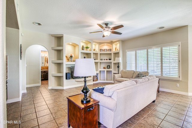 tiled living room with ceiling fan, arched walkways, a textured ceiling, built in features, and baseboards