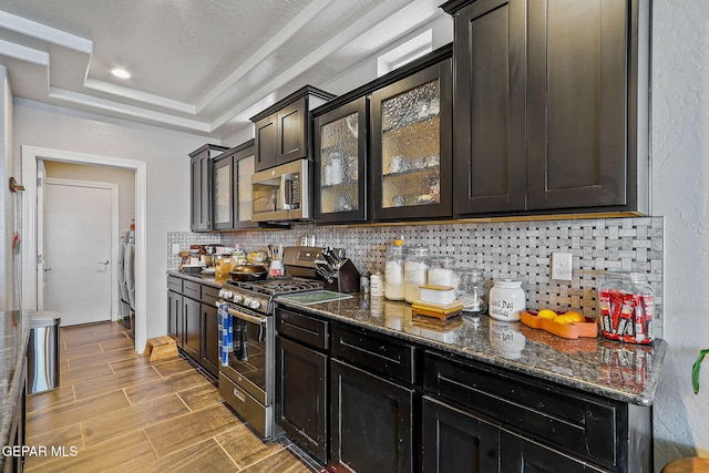 kitchen featuring stainless steel appliances, tasteful backsplash, light wood-type flooring, dark stone counters, and a tray ceiling