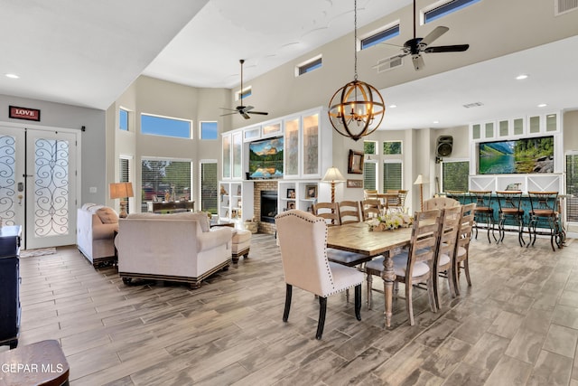 dining room featuring wood finished floors, ceiling fan with notable chandelier, a brick fireplace, visible vents, and a towering ceiling