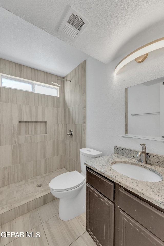 full bath with a textured ceiling, toilet, visible vents, tiled shower, and vanity
