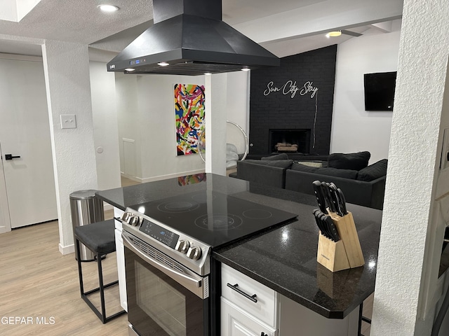 kitchen with wall chimney range hood, stainless steel range with electric cooktop, white cabinets, light wood-style flooring, and dark stone counters