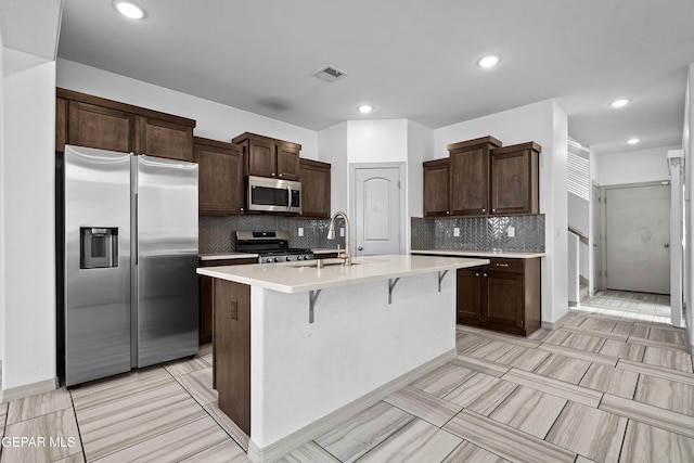 kitchen featuring dark brown cabinetry, appliances with stainless steel finishes, a sink, light countertops, and a kitchen island with sink