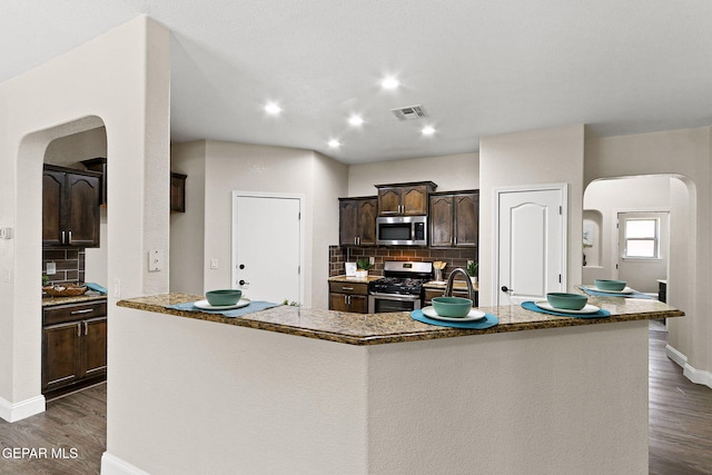 kitchen featuring decorative backsplash, dark brown cabinets, arched walkways, dark wood finished floors, and appliances with stainless steel finishes