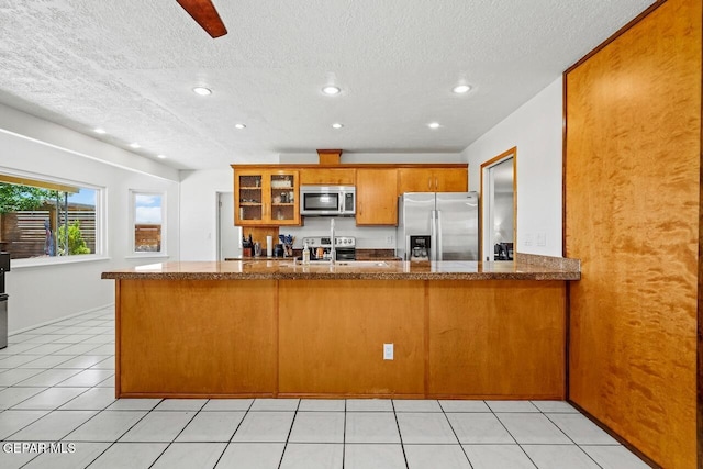 kitchen with stainless steel appliances, recessed lighting, brown cabinetry, glass insert cabinets, and a textured ceiling