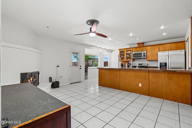 kitchen featuring stainless steel appliances, ceiling fan, a warm lit fireplace, brown cabinets, and glass insert cabinets