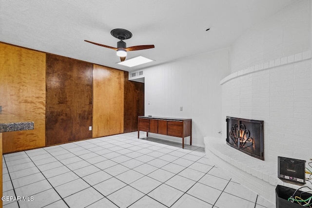 unfurnished living room with a ceiling fan, a brick fireplace, a skylight, light tile patterned floors, and wooden walls