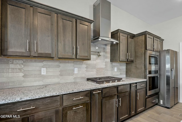 kitchen with wall chimney range hood, dark brown cabinets, light stone countertops, light wood finished floors, and stainless steel appliances