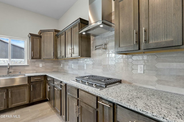 kitchen with wall chimney range hood, light stone counters, a sink, decorative backsplash, and stainless steel gas stovetop