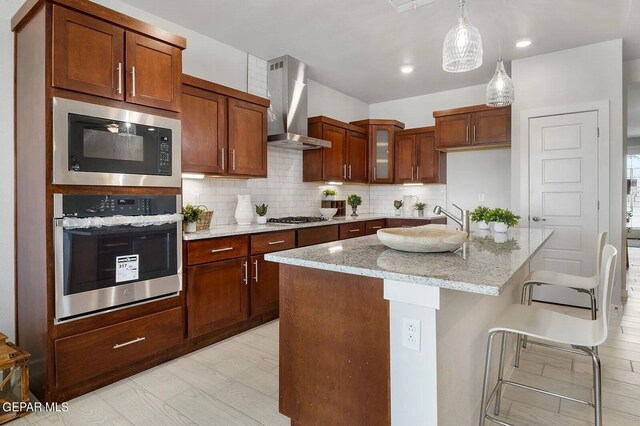 kitchen featuring wall chimney range hood, light stone countertops, stainless steel appliances, backsplash, and a center island with sink
