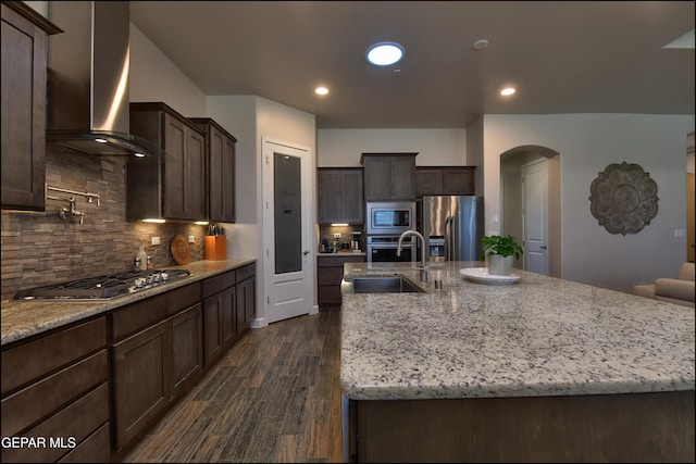 kitchen featuring wall chimney range hood, dark brown cabinetry, appliances with stainless steel finishes, a sink, and arched walkways