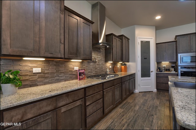 kitchen with dark brown cabinets, wall chimney range hood, tasteful backsplash, light stone counters, and appliances with stainless steel finishes