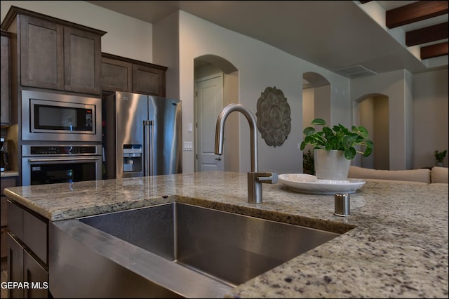 kitchen featuring dark brown cabinetry, appliances with stainless steel finishes, light stone countertops, a sink, and arched walkways