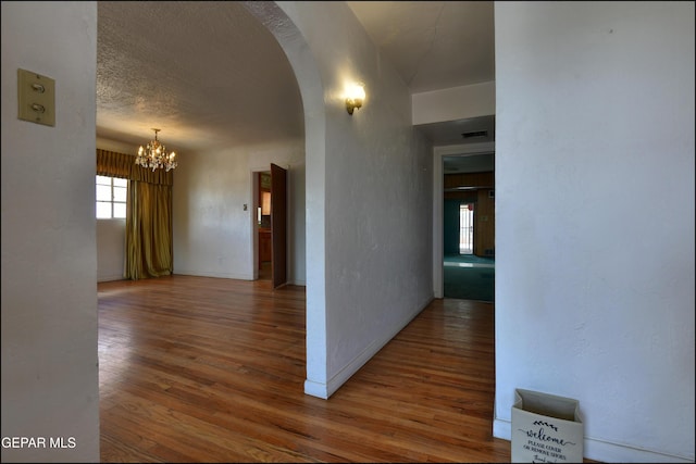 hall featuring wood finished floors, arched walkways, a chandelier, a textured wall, and baseboards