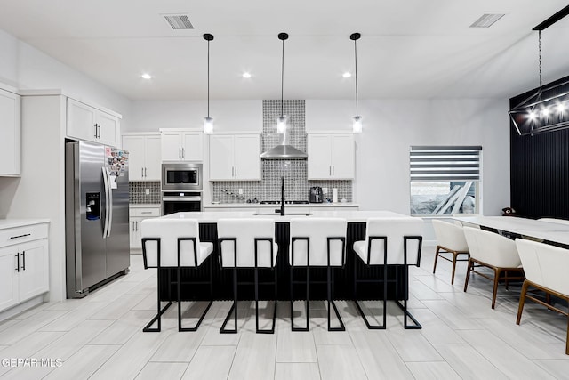 kitchen featuring backsplash, light countertops, stainless steel appliances, wall chimney exhaust hood, and an island with sink