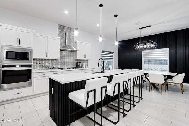 kitchen featuring wall chimney range hood, appliances with stainless steel finishes, a breakfast bar, light countertops, and a center island with sink