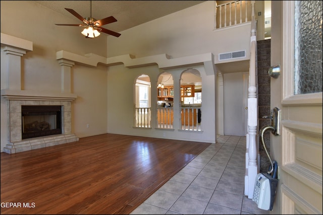 unfurnished living room with a tile fireplace, wood finished floors, a ceiling fan, visible vents, and a towering ceiling