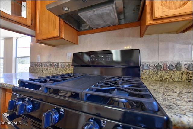 kitchen with brown cabinetry, range hood, light stone counters, gas stove, and decorative backsplash