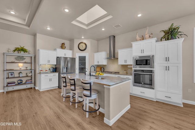 kitchen featuring a kitchen bar, appliances with stainless steel finishes, tasteful backsplash, light wood finished floors, and wall chimney exhaust hood