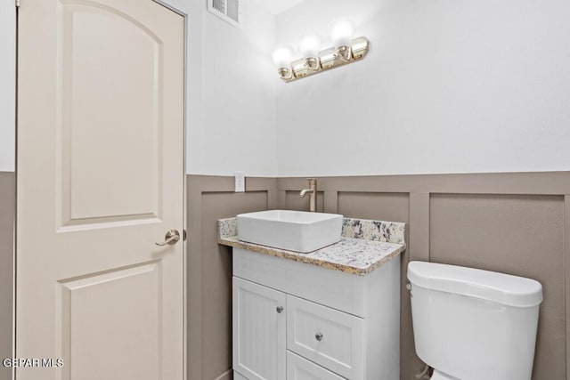 half bath featuring vanity, wainscoting, toilet, and a decorative wall