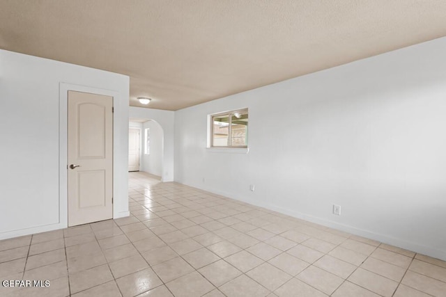 empty room featuring arched walkways, light tile patterned flooring, a textured ceiling, and baseboards