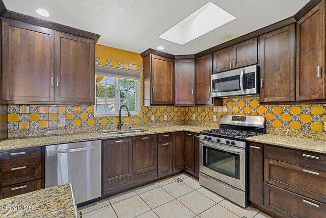 kitchen with stainless steel appliances, a sink, a skylight, light stone counters, and decorative backsplash