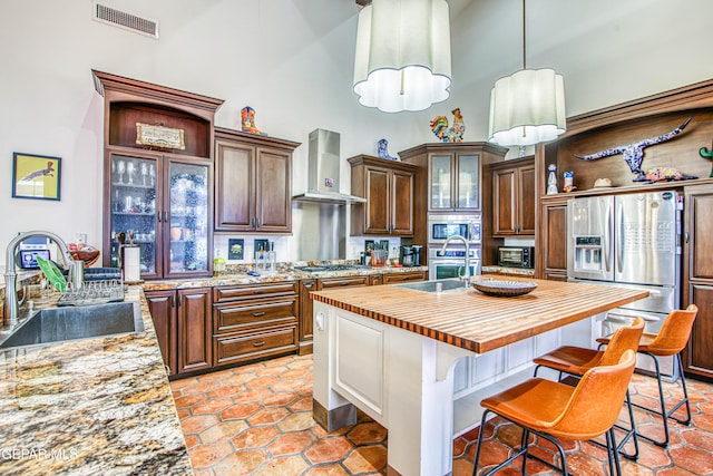 kitchen with wall chimney exhaust hood, appliances with stainless steel finishes, a sink, a towering ceiling, and butcher block countertops