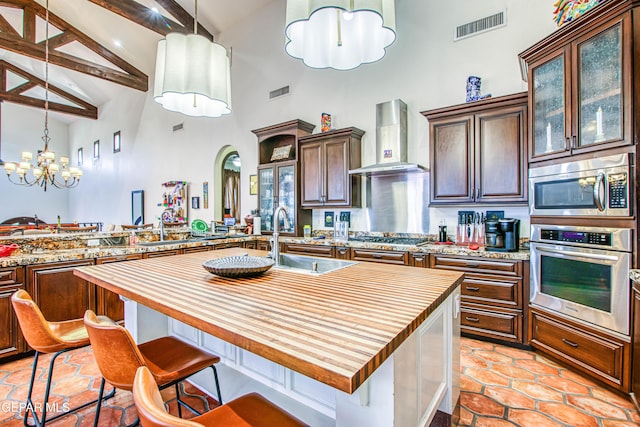 kitchen with appliances with stainless steel finishes, wall chimney range hood, butcher block countertops, high vaulted ceiling, and a sink