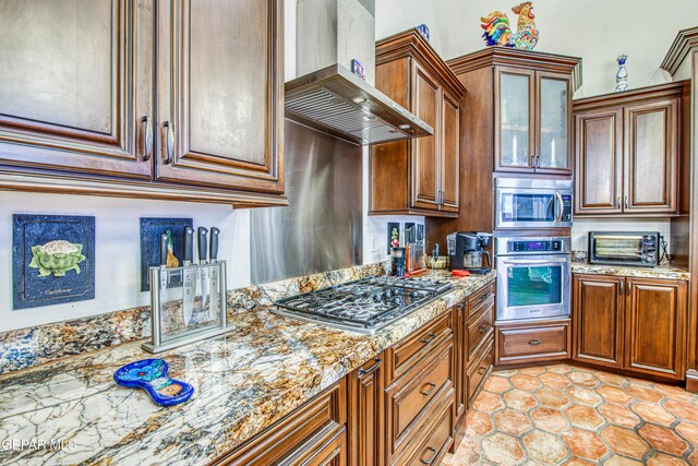 kitchen with appliances with stainless steel finishes, wall chimney range hood, light stone counters, and brown cabinets