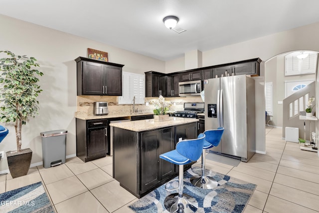 kitchen with stainless steel appliances, backsplash, a kitchen island, light tile patterned flooring, and light stone countertops
