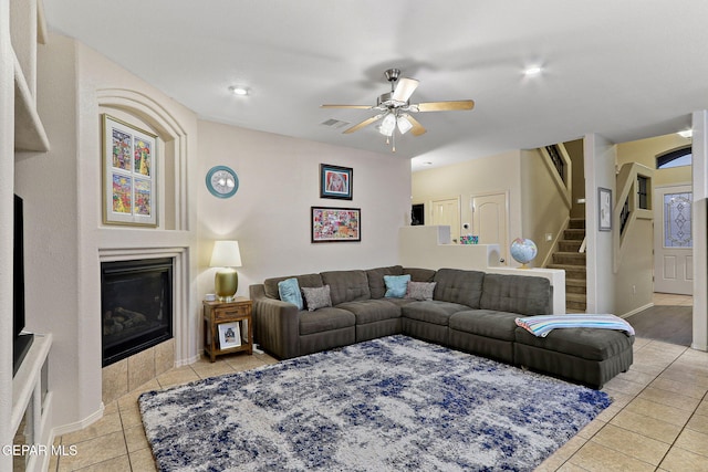 living area featuring a ceiling fan, light tile patterned floors, a tile fireplace, stairs, and baseboards