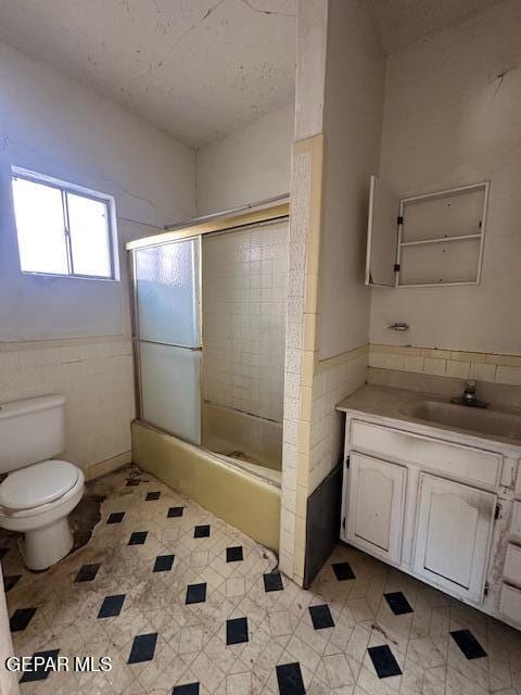 bathroom featuring tile walls, toilet, vanity, bath / shower combo with glass door, and a wainscoted wall