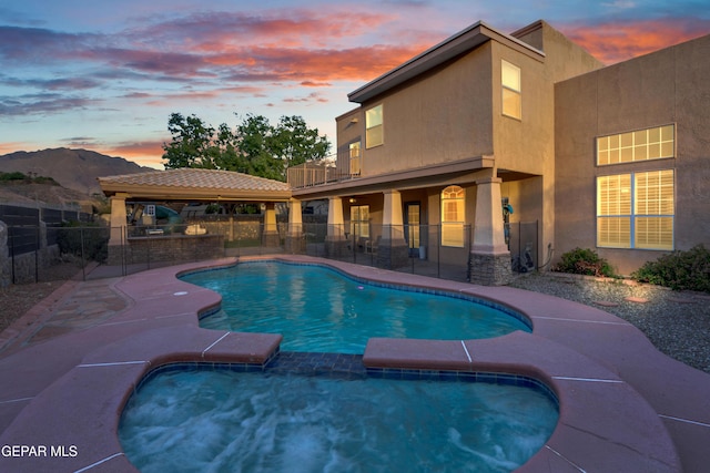 pool at dusk with fence, a patio area, a pool with connected hot tub, and a gazebo