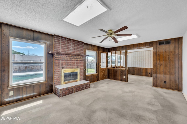 unfurnished living room featuring wooden walls, a textured ceiling, a ceiling fan, a brick fireplace, and carpet