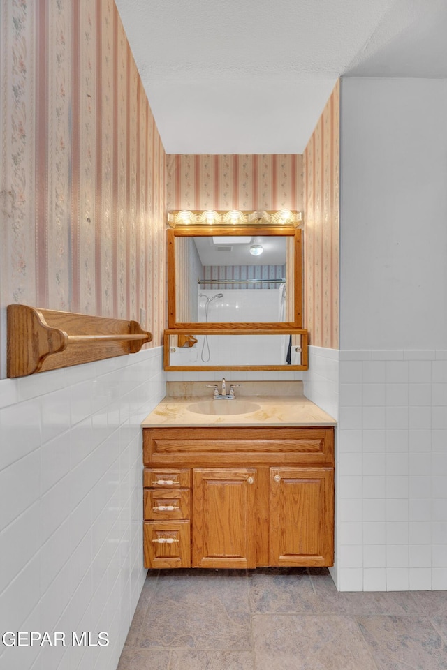 bathroom with wallpapered walls, vanity, wainscoting, and tile walls
