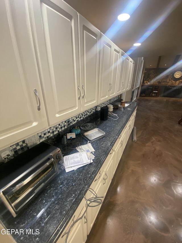 kitchen view of finished concrete floors, white cabinetry, decorative backsplash, and dark stone countertops