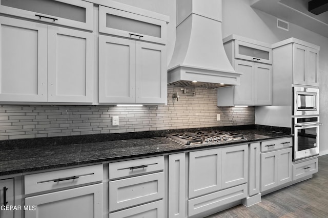 kitchen with appliances with stainless steel finishes, custom range hood, light wood-style floors, tasteful backsplash, and glass insert cabinets