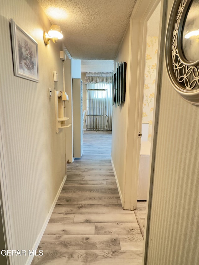 hall with light wood-type flooring, a textured ceiling, wallpapered walls, and baseboards