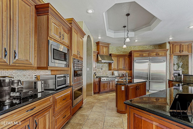 6664 Tuscany Ridge Dr, El Paso, TX 79912 - kitchen with built in appliances, an island with sink, backsplash, brown cabinets, and range hood