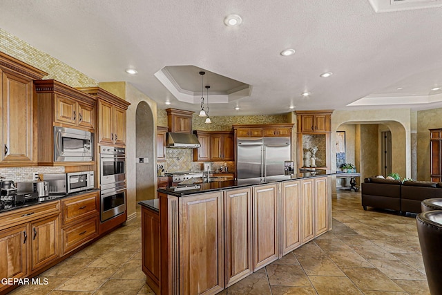 6664 Tuscany Ridge Dr, El Paso, TX 79912 - kitchen featuring a tray ceiling, built in appliances, arched walkways, dark countertops, and range hood