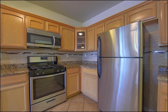 kitchen featuring stainless steel appliances, glass insert cabinets, tasteful backsplash, light tile patterned floors, and dark stone countertops