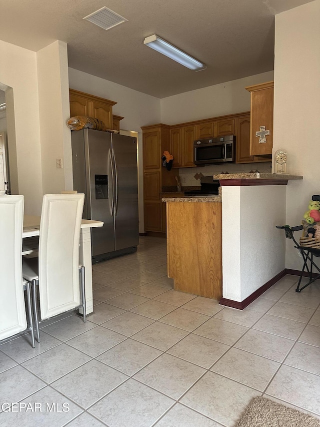 14045 Lago Seco Dr, Horizon City, TX 79928 - kitchen with stainless steel appliances, a peninsula, light tile patterned floors, and brown cabinetry